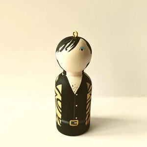 Puede incluir: Un adorno de muñeca de madera pintado a mano con cabello negro, una camisa negra con detalles dorados y un cinturón negro. La muñeca tiene una cara blanca con un ojo azul.
