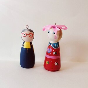 Op de afbeelding: Twee houten peg dolls. De ene pop draagt een marineblauwe outfit met een gele kraag en een rode bril. De andere pop draagt een rode jurk met witte sterren en een roze strik op haar hoofd. Beide poppen hebben metalen lussen om op te hangen.