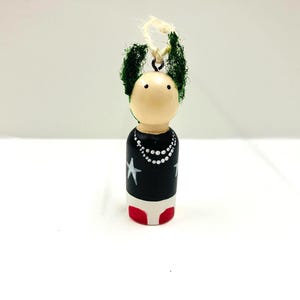 Puede incluir: Adorno de muñeca de madera con tono de piel claro, cabello verde y una camisa negra adornada con estrellas blancas y un collar de perlas. La base de la muñeca es blanca con una parte inferior roja y tiene una cuerda para colgar.
