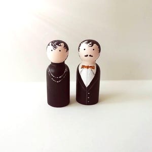 Può includere: Un paio di bambole di legno dipinte a mano vestite con abiti da sera neri. La donna indossa una collana e l'uomo un papillon.