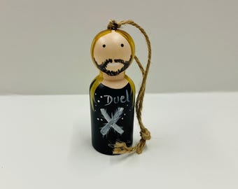 May include: A hand-painted wooden ornament depicting a person with long blonde hair, a black shirt with the word "Duel" and a white X on it, and a brown beard. The ornament has a brown twine loop for hanging.