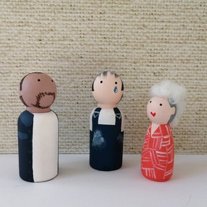 May include: Three hand-painted wooden peg dolls, each with a unique design. The doll on the left is a black man wearing a white shirt and a dark blue jacket. The middle doll is a man wearing a blue shirt and overalls. The doll on the right is a woman with white hair and a red and white patterned dress.