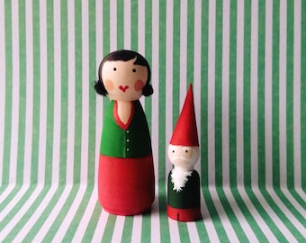 May include: A wooden doll couple, a woman with black hair and a red dress and a man with a red hat and a green beard. The dolls are standing on a green and white striped background.