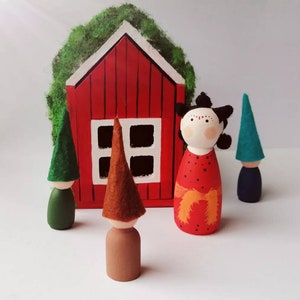 May include: A whimsical wooden scene featuring a red house with a green moss roof and several wooden figures. The figures include gnomes with felt hats and a woman in a red dress. The scene is set against a white background.