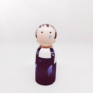 Puede incluir: Una muñeca de madera pintada a mano con cabello castaño, una camisa blanca y un mono azul. La muñeca tiene una cara pintada con una sola lágrima azul.