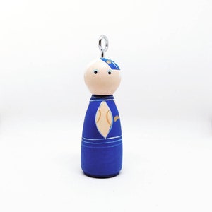 May include: A blue and white wooden doll ornament with a gold design. The doll has a white face with blue eyes and a blue and white hat. The doll is hanging from a silver loop.