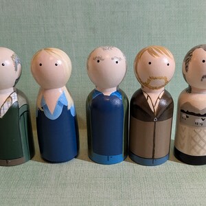 Könnte beinhalten: Fünf Holzpuppen mit bemalten Gesichtern und Kleidung. Die Puppen haben unterschiedliche Frisuren und Outfits, darunter ein grüner Mantel, ein blaues Kleid und ein brauner Anzug. Jede Puppe ist etwa 10 cm groß.