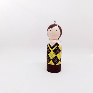 May include: A wooden doll ornament with brown hair, a white collar, and a black and yellow checked sweater.