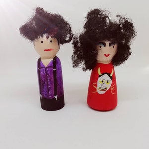 Pode incluir: Duas bonecas de madeira com cabelo de lã preto. A boneca à esquerda está vestindo uma camisa roxa com suspensórios pretos e calças pretas. A boneca à direita está vestindo uma camisa vermelha com um desenho de máscara amarela e branca.