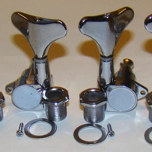May include: Four chrome-plated bass guitar tuning keys with white plastic buttons. Each key has a three-pronged tuning peg and a threaded shaft. The keys are arranged in a row, with the tuning pegs facing the viewer. The keys are accompanied by four washers and four screws.