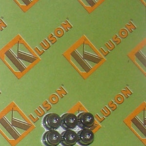 May include: A close-up of six silver-colored metal furniture feet. The feet are arranged in a staggered pattern on a green background with a repeating logo that says "KLUSON".