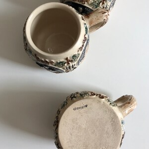 May include: Three ceramic beer steins with intricate relief designs. The steins feature a beige base color with detailed floral and vine patterns in shades of green, blue, and brown. The bottom of one stein is marked "GERMANY" and "2020".