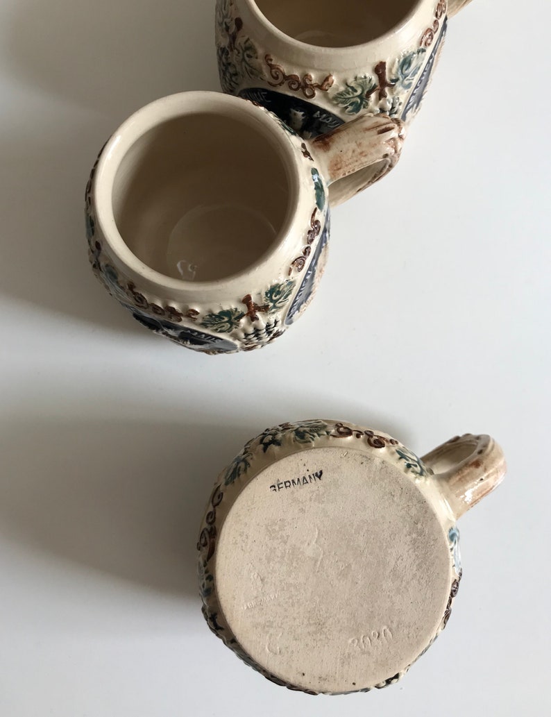 May include: Three ceramic beer steins with intricate relief designs. The steins feature a beige base color with detailed floral and vine patterns in shades of green, blue, and brown. The bottom of one stein is marked "GERMANY" and "2020".