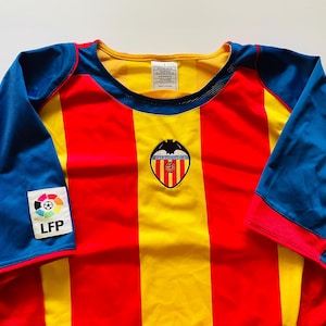 May include: A short-sleeved sports jersey featuring vertical red and yellow stripes on the front. The sleeves are blue with red trim. A Valencia CF team crest is centered on the chest, and an LFP league patch is visible on the right sleeve.