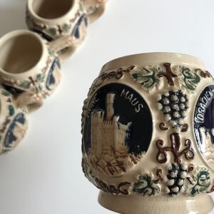 May include: A collection of small, beige ceramic mugs with detailed relief designs. Each mug features a dark blue circular emblem with a castle illustration and the word "MAUS". The mugs are decorated with grape vines and other embossed details.