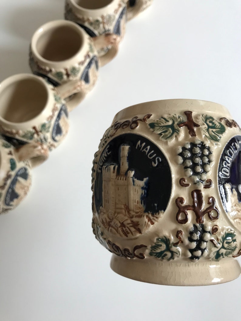 May include: A collection of small, beige ceramic mugs with detailed relief designs. Each mug features a dark blue circular emblem with a castle illustration and the word "MAUS". The mugs are decorated with grape vines and other embossed details.