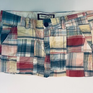 May include: A plaid patchwork mini skirt featuring a multi-colored pattern of red, blue, yellow, and white squares. The skirt has a button closure, belt loops, and a visible Abercrombie & Fitch label on the interior waistband.