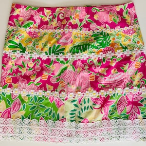 Op de afbeelding: Een roze en groene bloemenrok met witte kant. De rok heeft een veelkleurig bloemenpatroon met een roze achtergrond. De rok heeft een witte kant rond de zoom.