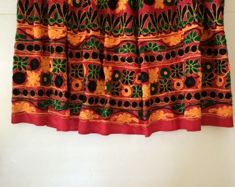 May include: Close-up of a red fabric with intricate embroidery. The design features rows of floral and geometric patterns in yellow, green, and black thread. The fabric has a textured appearance, with a scalloped edge.