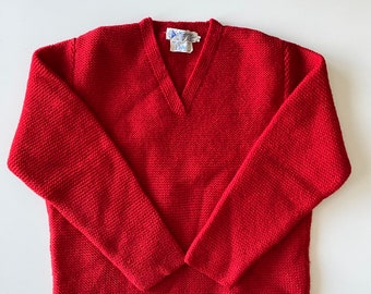 May include: A red knit sweater with a V-neck. The sweater has long sleeves and a textured knit pattern. A small tag is visible near the neckline. The sweater is displayed on a white surface.