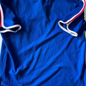 以下が含まれることがあります： ロイヤルブルーの半袖ポロシャツで、襟には白いトリムが施されています。袖には白いパイピングで縁取られた赤いストライプのアクセントがあります。シャツはストレートな裾で、下端に沿って白いトリムのディテールがあしらわれています。