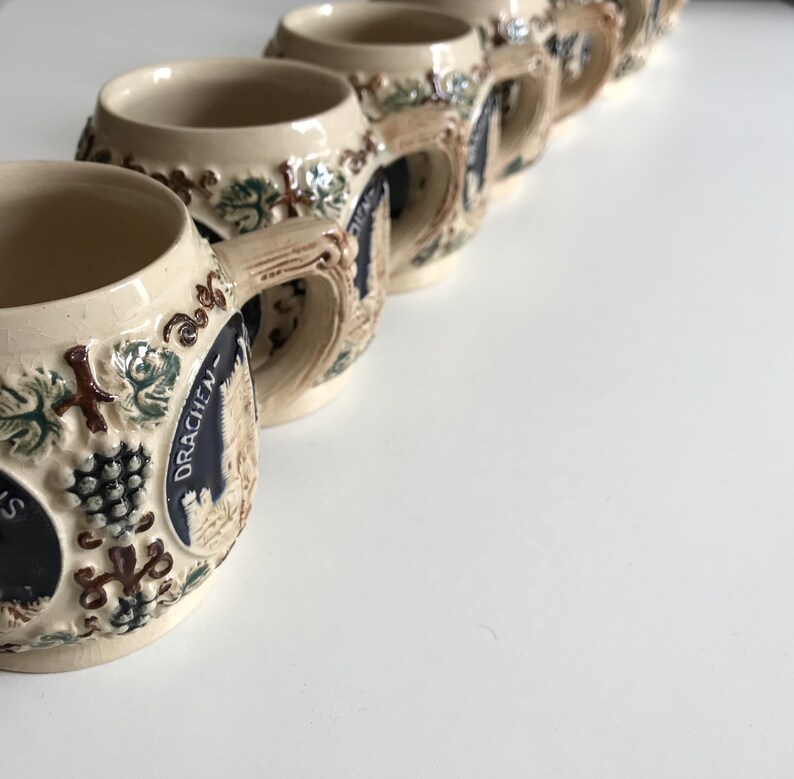 May include: A row of beige ceramic beer steins with ornate relief designs. Each stein features a handle, a dark blue emblem with text, and grape and leaf motifs. The steins are arranged diagonally on a white surface.