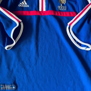 May include: A blue Adidas polo shirt featuring a red horizontal stripe across the chest, white trim on the sleeves and collar, and the FFF logo with a gold star on the left chest. An authentic licensed product tag is visible at the bottom hem.