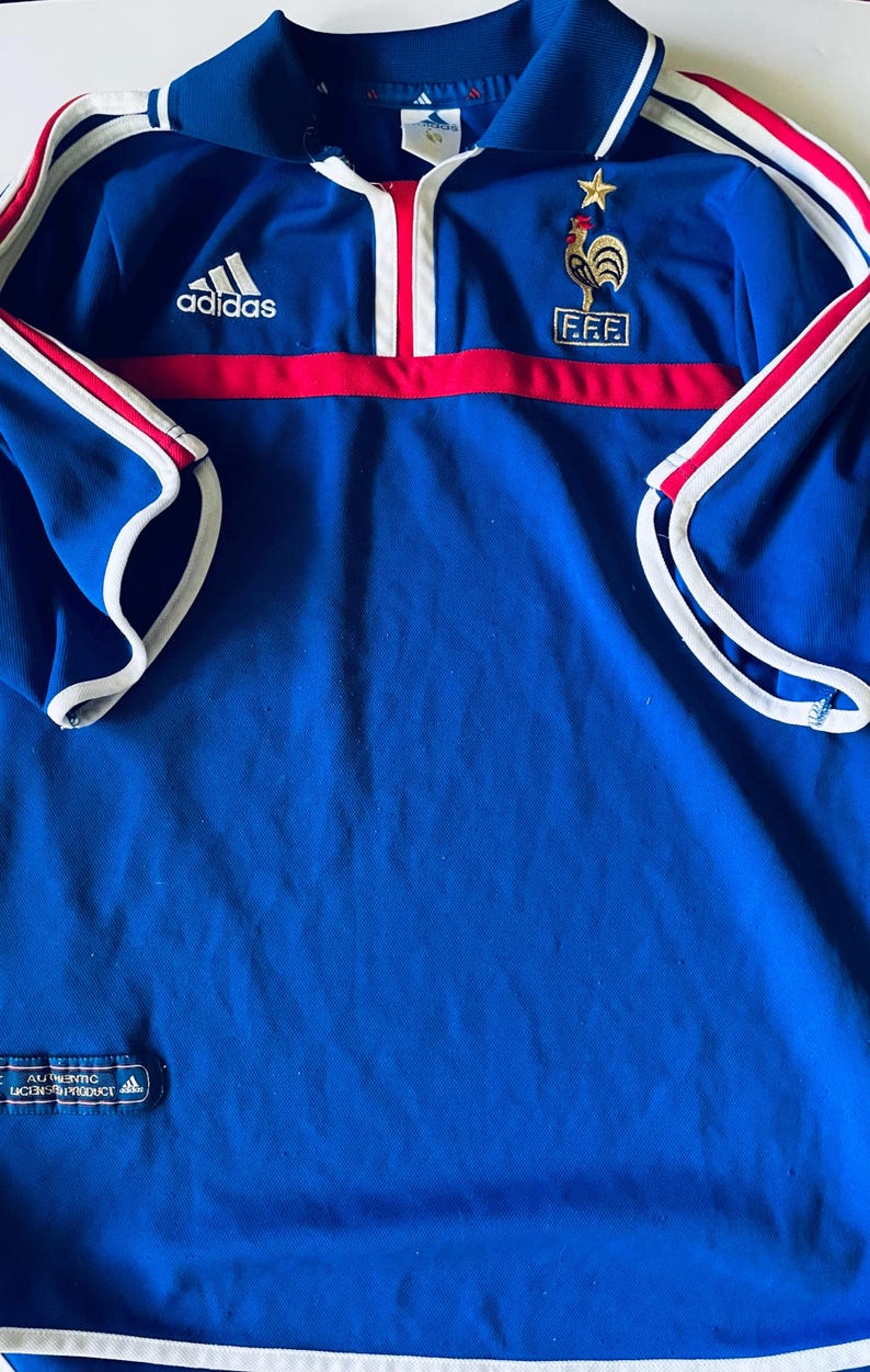 May include: A blue Adidas polo shirt featuring a red horizontal stripe across the chest, white trim on the sleeves and collar, and the FFF logo with a gold star on the left chest. An authentic licensed product tag is visible at the bottom hem.