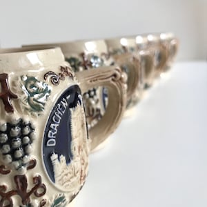 May include: A row of vintage ceramic beer steins with ornate handles. Each stein features a relief design of grapes and leaves, with a central emblem displaying the word "DRACHEN" and a castle illustration. The steins have a cream-colored base with brown, blue, and green accents.