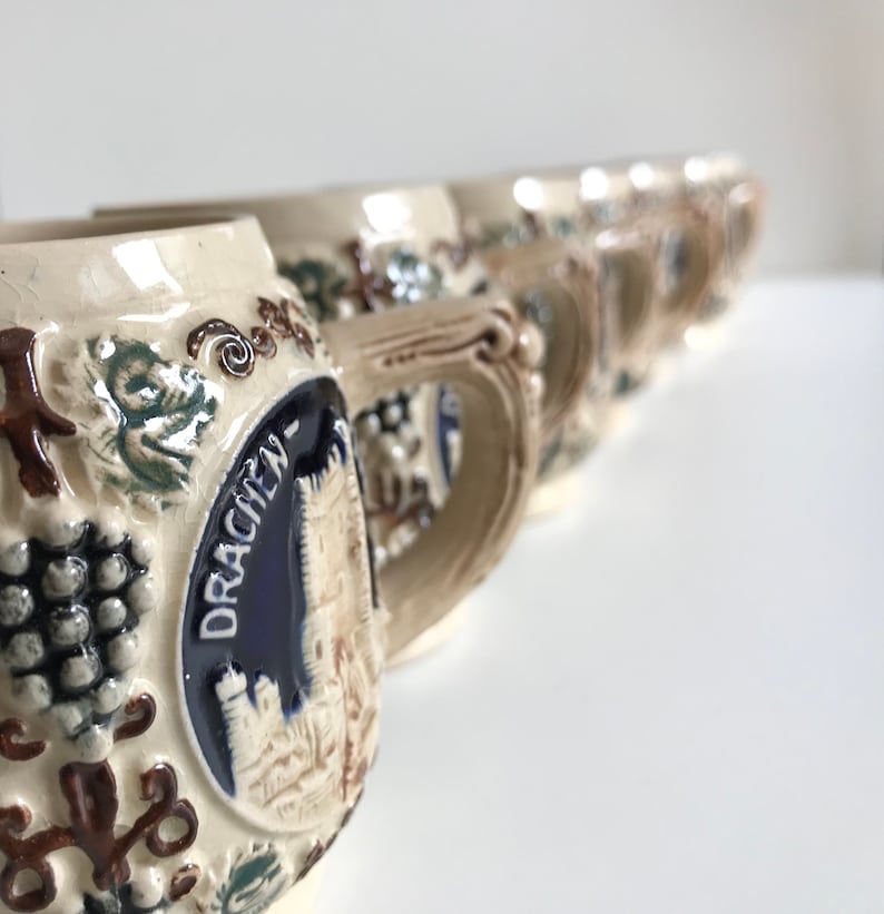 May include: A row of vintage ceramic beer steins with ornate handles. Each stein features a relief design of grapes and leaves, with a central emblem displaying the word "DRACHEN" and a castle illustration. The steins have a cream-colored base with brown, blue, and green accents.