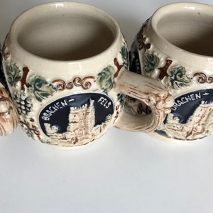 May include: A collection of beige ceramic beer steins with detailed relief designs. Each stein features a castle emblem with the words "DRACHEN-FELS" and is adorned with grapevines and leaves. The handles are textured to resemble wood.