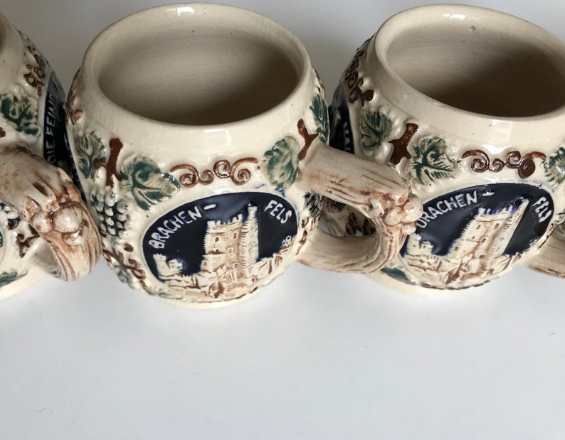 May include: A collection of beige ceramic beer steins with detailed relief designs. Each stein features a castle emblem with the words "DRACHEN-FELS" and is adorned with grapevines and leaves. The handles are textured to resemble wood.