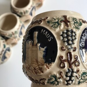 May include: A set of small, cream-colored ceramic shot glasses with embossed designs. Each glass features a detailed illustration of a castle, grapes, and foliage, with dark blue and brown accents. The glasses have a slightly aged, vintage appearance.