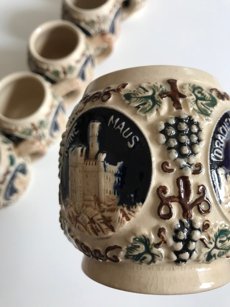 May include: A set of small, cream-colored ceramic shot glasses with embossed designs. Each glass features a detailed illustration of a castle, grapes, and foliage, with dark blue and brown accents. The glasses have a slightly aged, vintage appearance.