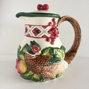 Może przedstawiać: Biały dzbanek ceramiczny z zieloną pokrywką i brązowym uchwytem. Dzbanek jest ozdobiony bażantem, liśćmi i czerwonymi jagodami.