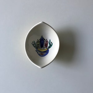 以下が含まれることがあります： 葉の形をした白い陶器の皿。手描きで、カラフルな家が3つ、緑の木々、青い空が描かれている。