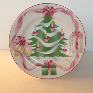 Op de afbeelding: Een wit keramiek kerstbord met een groene kerstboom, roze linten en cadeaus. De boom is versierd met witte kaarsen en roze harten.