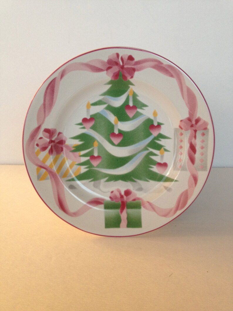 Op de afbeelding: Een wit keramiek kerstbord met een groene kerstboom, roze linten en cadeaus. De boom is versierd met witte kaarsen en roze harten.