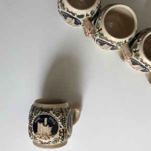 May include: A collection of small, beige ceramic beer steins. Each stein features a detailed illustration of a castle within a dark blue circle, surrounded by grape vines. The steins have small handles and are arranged on a white surface.