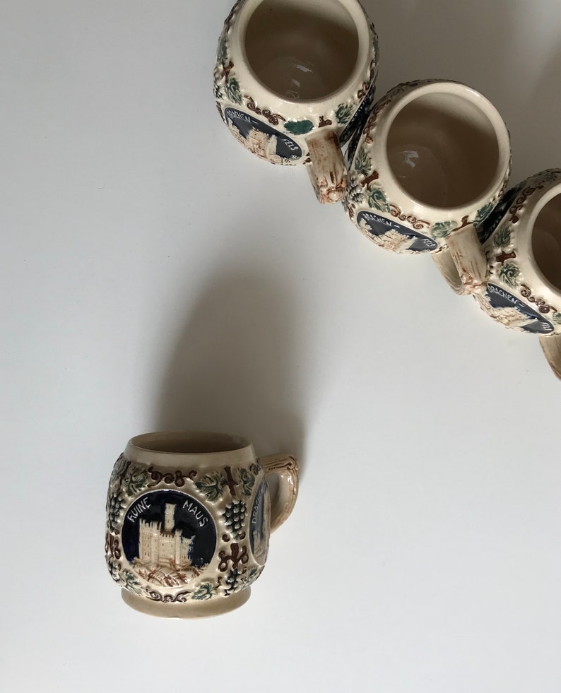 May include: A collection of small, beige ceramic beer steins. Each stein features a detailed illustration of a castle within a dark blue circle, surrounded by grape vines. The steins have small handles and are arranged on a white surface.