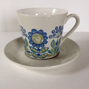 Może przedstawiać: Biały ceramiczny kubek i spodek z niebieskim i zielonym wzorem kwiatowym. Kubek ma szeroką, zaokrągloną podstawę i uchwyt. Spodek jest okrągły i ma lekko uniesiony brzeg.