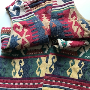 Può includere: Un gilet tessuto con un motivo geometrico colorato nei toni del rosso, blu, verde e giallo. Il gilet ha un orlo con frange e una fodera beige. Il design presenta forme e linee ripetute, creando un aspetto strutturato.