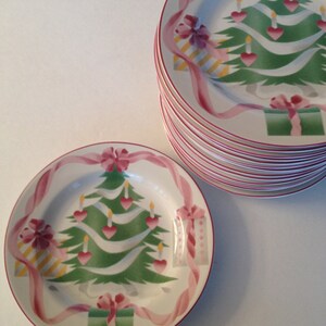 Op de afbeelding: Een set witte keramische kerstplaten met een groen kerstboomontwerp. De boom is versierd met roze harten, gele kaarsen en roze linten. De platen hebben een rode rand.
