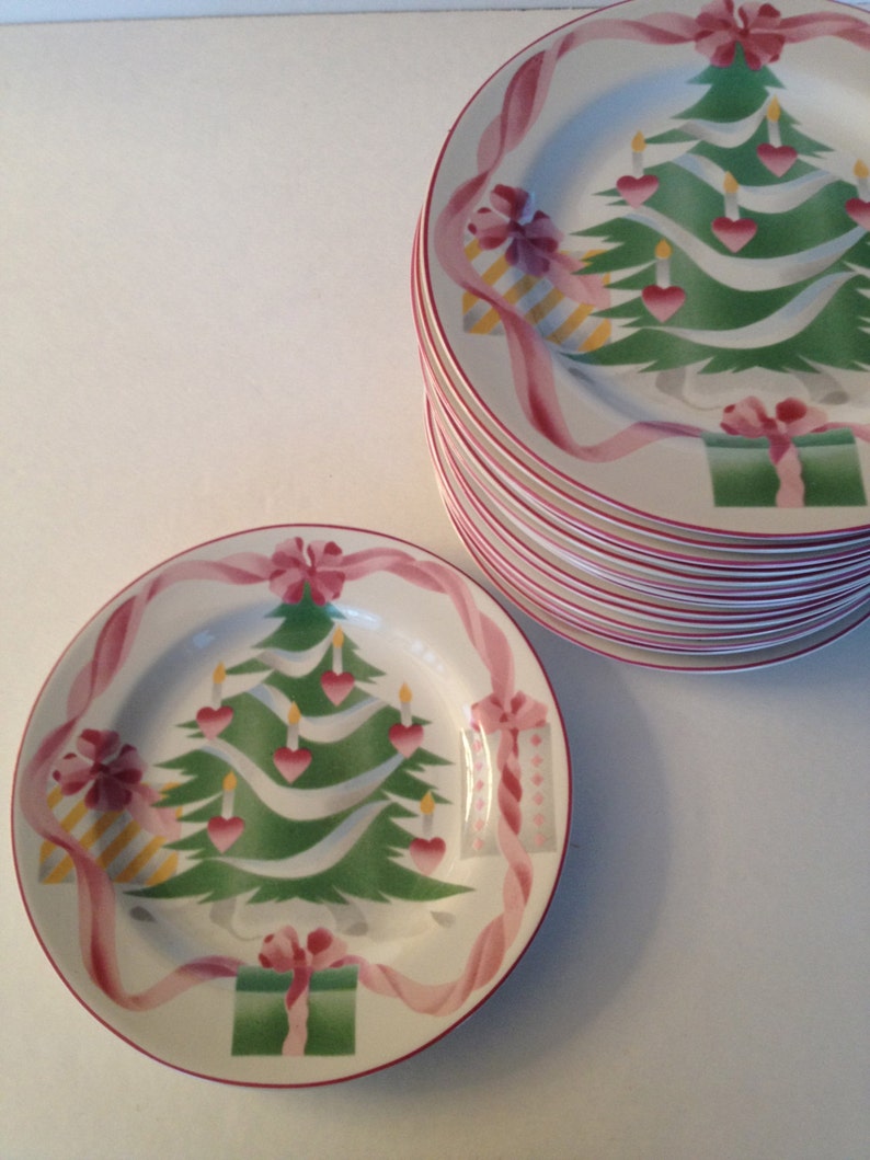 Op de afbeelding: Een set witte keramische kerstplaten met een groen kerstboomontwerp. De boom is versierd met roze harten, gele kaarsen en roze linten. De platen hebben een rode rand.