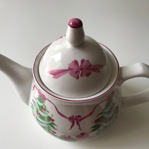 Puede incluir: Tetera de cerámica blanca con un diseño de árbol de Navidad rosa y verde. La tetera tiene una tapa con un lazo rosa.