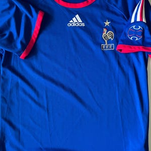 May include: A blue Adidas short-sleeve jersey featuring a red crew neckline and sleeve trim. The shirt displays a white Adidas logo, a gold star, and a rooster emblem above the text FFF. White stripes accent the shoulders, and a Climalite logo is visible.