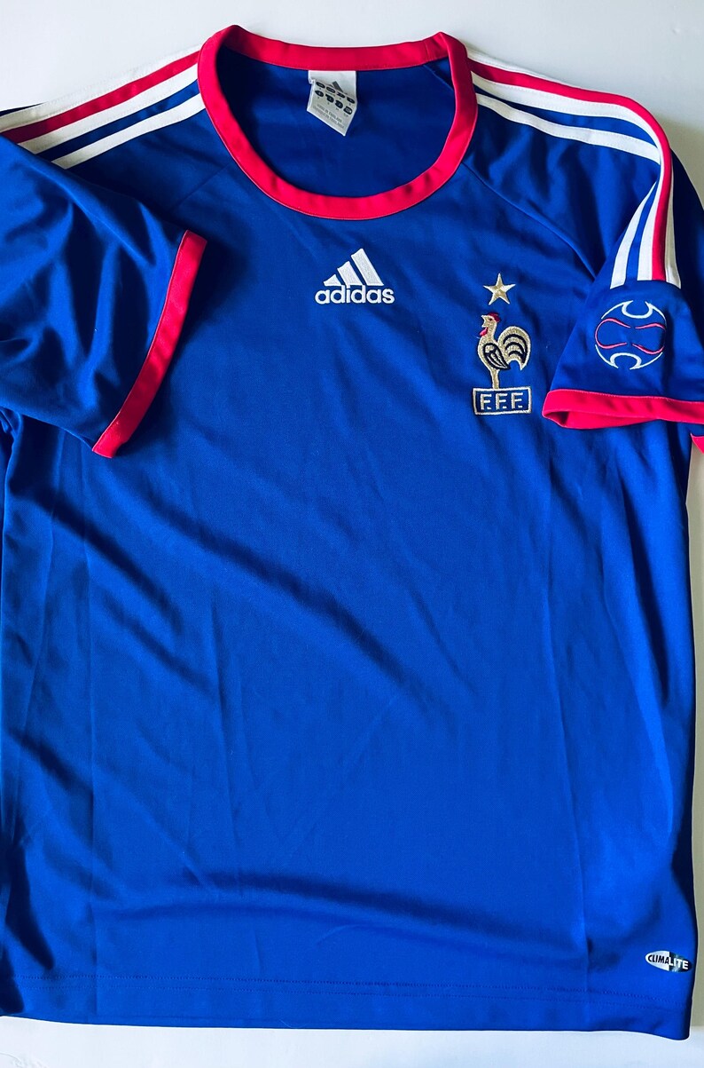 May include: A blue Adidas short-sleeve jersey featuring a red crew neckline and sleeve trim. The shirt displays a white Adidas logo, a gold star, and a rooster emblem above the text FFF. White stripes accent the shoulders, and a Climalite logo is visible.