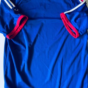May include: A short-sleeved blue athletic jersey featuring a red crew neckline and red sleeve cuffs. The shoulders are accented with two white stripes that run down the length of the sleeves. A small dark logo is visible on the right sleeve.