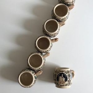 May include: A collection of ceramic beer steins with ornate handles and detailed relief designs. The steins are beige with blue, green, and brown accents. One stein features a castle emblem and the text "Rheine Maus".