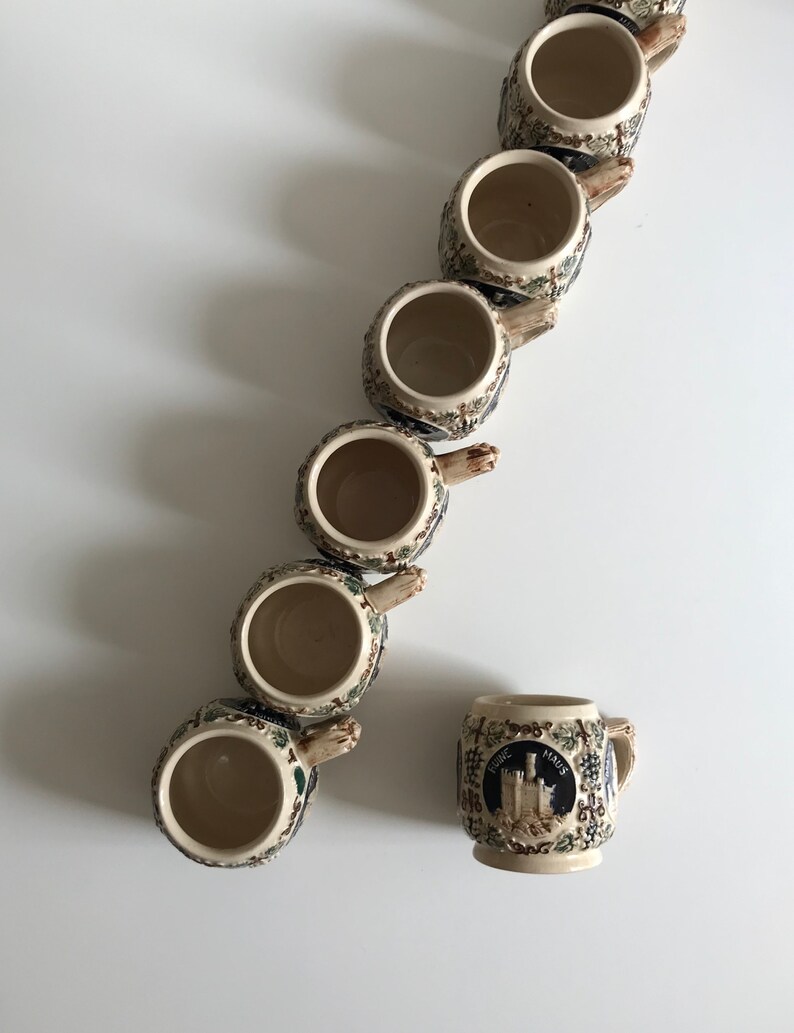 May include: A collection of ceramic beer steins with ornate handles and detailed relief designs. The steins are beige with blue, green, and brown accents. One stein features a castle emblem and the text "Rheine Maus".